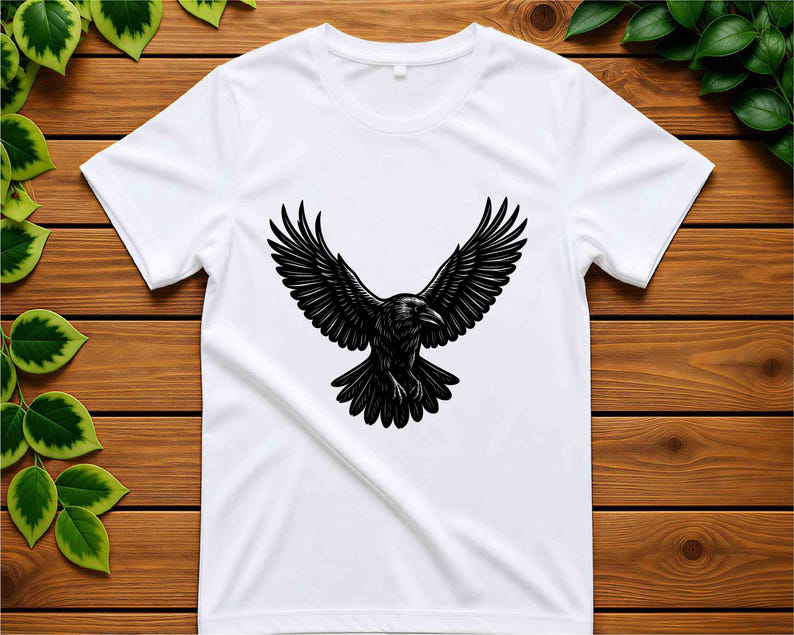May include: A white short-sleeved t-shirt featuring a centered black graphic illustration of a raven with its wings spread wide in flight. The bird is depicted with detailed feather textures and a sharp beak, positioned against the plain white fabric.