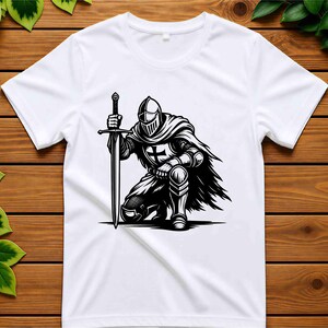 May include: A white short-sleeved t-shirt featuring a black and white graphic print of a knight in full armour kneeling while holding a long sword. The knight wears a helmet and a cape, with a cross emblem visible on the chest plate of the armour.