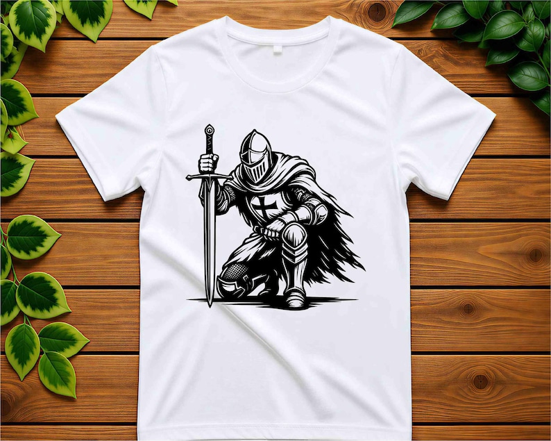 May include: A white short-sleeved t-shirt featuring a black and white graphic print of a knight in full armour kneeling while holding a long sword. The knight wears a helmet and a cape, with a cross emblem visible on the chest plate of the armour.