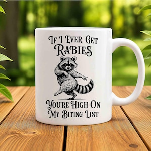 May include: A white ceramic mug featuring a black ink illustration of a dancing raccoon in the center. The text above the raccoon reads If I Ever Get Rabies and the text below the raccoon reads You're High On My Biting List in a bold font.