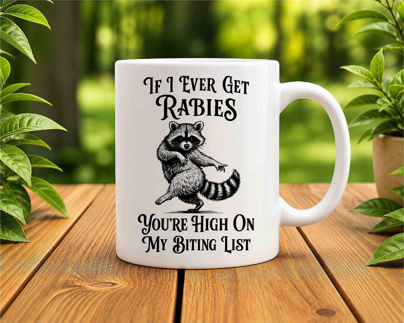 May include: A white ceramic mug featuring a black ink illustration of a dancing raccoon in the center. The text above the raccoon reads If I Ever Get Rabies and the text below the raccoon reads You're High On My Biting List in a bold font.