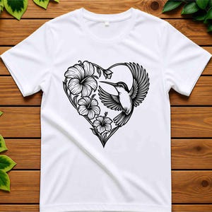 K&ouml;nnte beinhalten: Ein wei&szlig;es T-Shirt mit kurzen &Auml;rmeln und einer schwarzen Strichgrafik auf der Brust. Das Design zeigt einen fliegenden Kolibri zusammen mit drei Hibiskusbl&uuml;ten, die zusammen die Form eines Herzens bilden.