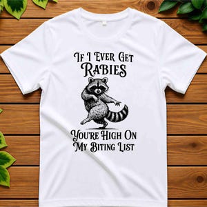 May include: A white short-sleeved t-shirt featuring a black graphic illustration of a dancing raccoon in the center. The text above the raccoon reads "If I Ever Get Rabies" and the text below the raccoon reads "You're High On My Biting List."