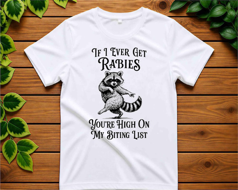 May include: A white short-sleeved t-shirt featuring a black graphic illustration of a dancing raccoon in the center. The text above the raccoon reads "If I Ever Get Rabies" and the text below the raccoon reads "You're High On My Biting List."