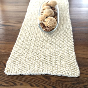 May include: Cream-colored, rectangular table runner with a textured, knitted appearance. A clear, oval-shaped glass dish filled with decorative wooden flowers and potpourri sits atop the runner. The runner is displayed on a dark wood surface.