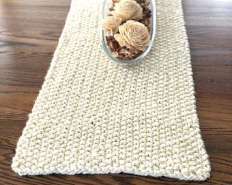 May include: Cream-colored, rectangular table runner with a textured, knitted appearance. A clear, oval-shaped glass dish filled with decorative wooden flowers and potpourri sits atop the runner. The runner is displayed on a dark wood surface.