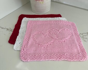 May include: Three knitted washcloths in red, white, and pink. The pink washcloth features two heart shapes. The washcloths are square with a textured border. A vase of pink flowers is in the background.