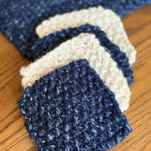 May include: A stack of handmade, square crochet coasters in shades of navy blue and cream. The coasters have a textured, nubby appearance, and are arranged on a wooden surface. The navy blue coasters have flecks of white.
