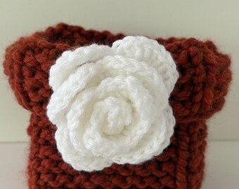 May include: A handmade, crochet container in a rich, reddish-brown hue. A large, white crochet rose is centered on the front. The container has a square shape with a slightly flared top, showcasing a textured stitch pattern.