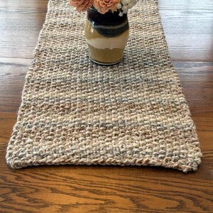 May include: A woven beige and brown table runner with a textured surface. A ceramic vase with a dark brown top, green stripe, and tan base holds peach-colored flowers and white filler flowers. The runner is on a wooden table.