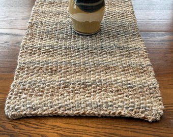 May include: A woven beige and brown table runner with a textured surface. A ceramic vase with a dark brown top, green stripe, and tan base holds peach-colored flowers and white filler flowers. The runner is on a wooden table.
