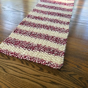May include: A rectangular, hand-knitted table runner with alternating stripes of cream and burgundy. The thick yarn creates a textured, cozy appearance. The runner is displayed on a wooden surface.