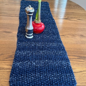 May include: A navy blue knitted table runner with white flecks, displayed on a wooden table. A silver pepper grinder and a red bulb with a green sprout are placed on the runner, creating a decorative centerpiece.