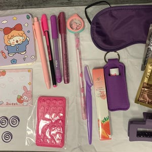 May include: A collection of pink and purple themed items including a small notebook, pens, a sleep mask, a lip mask, eye patches, and other small accessories.
