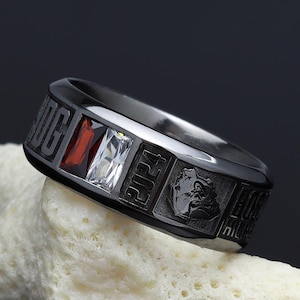 May include: A black ring with red and white gemstones and engraved details. The ring is displayed on a piece of white coral against a dark background. The ring has the year 2024 engraved on it.