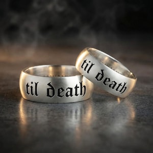 May include: Two silver rings with the phrase "til death" engraved in a gothic font. The rings are displayed on a dark, textured surface, with a blurred background.