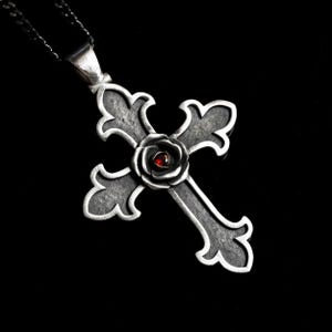 May include: A silver-toned pendant necklace featuring a gothic cross design. The cross has ornate edges and a rose in the center with a small, red gemstone. The necklace hangs on a dark chain against a black background.
