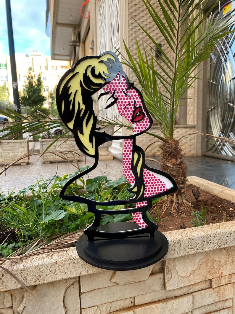 May include: A black metal sculpture of a woman's bust with a pop art style design. The woman's hair is yellow, her face is pink with white polka dots, and her lips are red. The sculpture is on a black base.