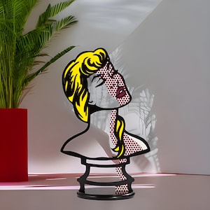 May include: A black metal sculpture of a woman's bust with a yellow hairdo and a red and white patterned face. The sculpture is on a black pedestal.