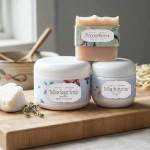 May include: A wooden cutting board displays a bar of strawberry tallow soap, two jars of Tallow Sugar Scrub and Tallow Moisturizer, and a piece of white soap. The jars have floral labels. A bowl and spoons are in the background.
