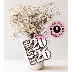 Può includere: Un vaso bianco con un bouquet di fiori bianchi. Un'etichetta con stampa leopardata con la scritta "Class of 2026" e un fiocco rosa è attaccata. È visibile anche un cerchio rosa con le parole "Instant Download".