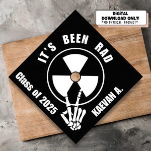 Op de afbeelding: Zwarte afstudeerhoed met witte tekst: "IT'S BEEN RAD", "Class of 2025" en de naam "KAEVAN A.". Een wit stralingssymbool en een skelet hand die een vredesteken maakt, zijn ook te zien. Alleen digitale download.