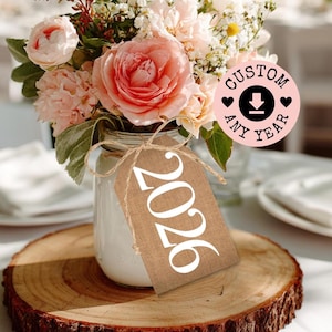 May include: A floral arrangement in a mason jar with a burlap tag that reads "2026". The flowers are a mix of pink and white blooms. The jar sits on a wooden slice. A pink circle with the words "CUSTOM ANY YEAR" is also visible.