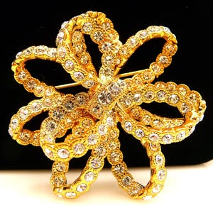 May include: A gold-toned brooch in the shape of a bow, encrusted with clear, sparkling rhinestones. The brooch has a pin on the back for secure fastening. The design is intricate and detailed, with a vintage aesthetic.