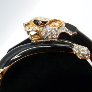 May include: A gold-toned and black enamel bracelet featuring a panther head with crystal accents. The panther's head is adorned with clear crystals and a black eye. The bracelet has a hinged design and a gold chain.