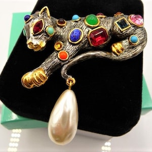 May include: A silver-toned metal brooch in the shape of a stylized leopard, adorned with colorful gemstones and gold accents. The brooch features a large, pear-shaped faux pearl pendant. The leopard is posed in a walking position.