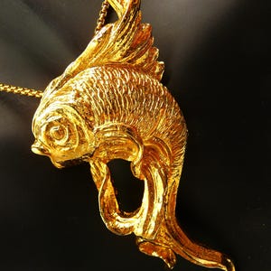 May include: A gold-colored fish pendant necklace. The pendant is a detailed sculpture of a fish with textured scales and flowing fins. The necklace chain is also gold-colored and delicate.