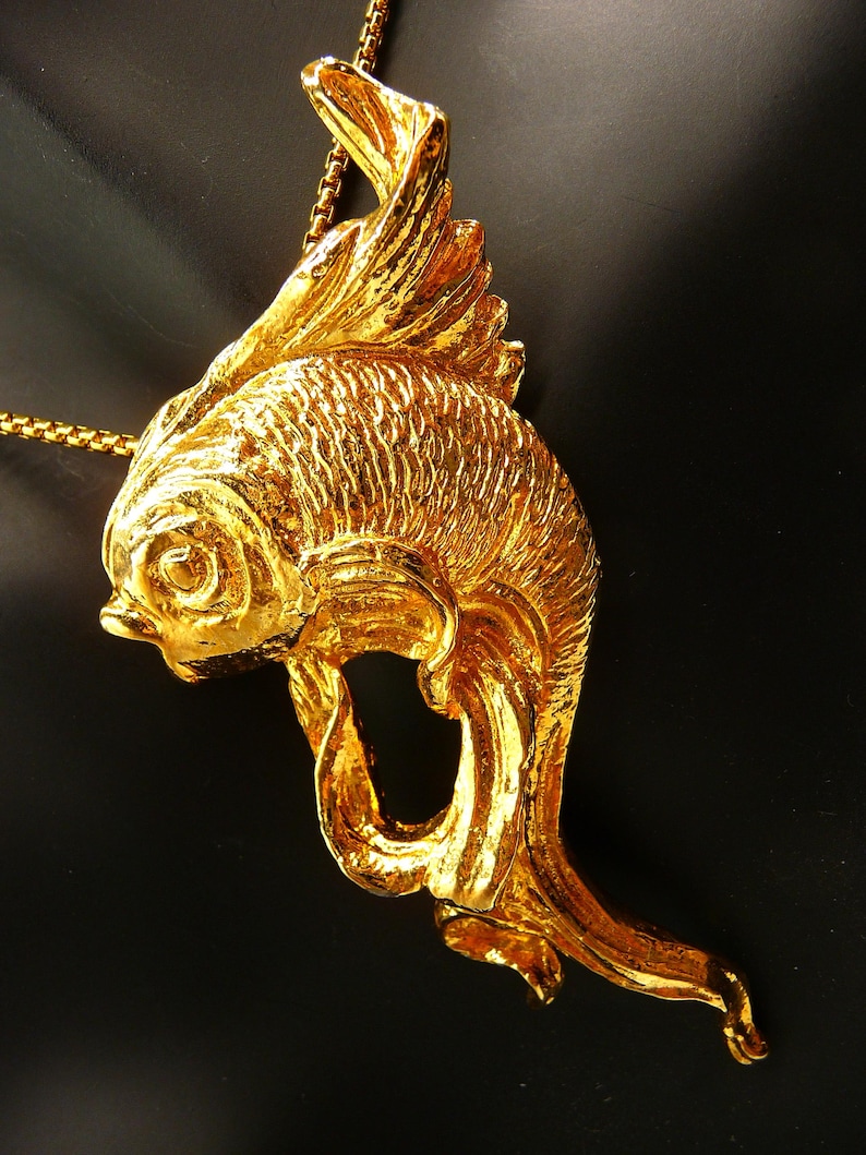 May include: A gold-colored fish pendant necklace. The pendant is a detailed sculpture of a fish with textured scales and flowing fins. The necklace chain is also gold-colored and delicate.