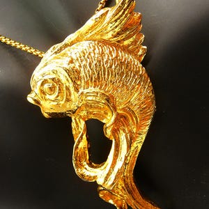 May include: A gold-colored fish pendant necklace. The pendant is a detailed sculpture of a fish with textured scales and flowing fins. The necklace chain is also gold-colored, creating a cohesive and elegant look. The fish pendant is a unique piece of jewelry.