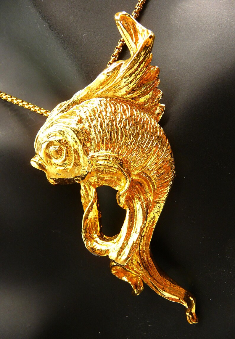 May include: A gold-colored fish pendant necklace. The pendant is a detailed sculpture of a fish with textured scales and flowing fins. The necklace chain is also gold-colored, creating a cohesive and elegant look. The fish pendant is a unique piece of jewelry.