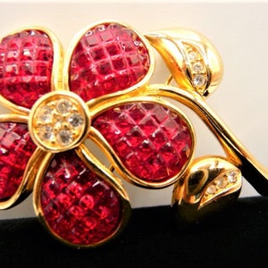 May include: A gold-toned flower brooch with five red, faceted petals. The center of the flower and the leaves have small, clear crystals. The brooch is a floral design, suitable for adding a touch of elegance to any outfit.