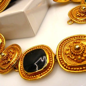 May include: Close-up of a gold-toned necklace with black enamel accents. The necklace features ornate, geometric links with a repeating pattern of raised details. The links are connected by small, gold-toned rings. The jewelry is displayed on a white surface.