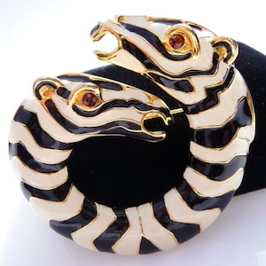 May include: A vintage brooch featuring a design of two stylized zebra heads in a circular arrangement. The brooch has a gold-tone metal base with black and white enamel stripes. The eyes are small, round, and red. A unique piece of jewelry.