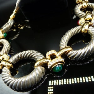 May include: A silver-toned necklace with a twisted rope design, featuring gold accents and colorful gemstones. The necklace has a series of large, circular links connected by smaller gold links. Gemstones include red, blue, and green stones.