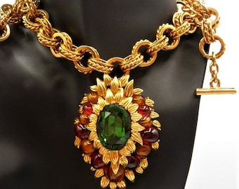 May include: A gold-tone chain necklace featuring a large, oval-shaped pendant. The pendant has a central faceted green stone surrounded by smaller, round, deep red and brown cabochon stones set in a textured, leaf-like gold metal frame.