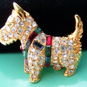May include: A gold-toned dog brooch encrusted with clear rhinestones. The dog has a collar of red, green, and blue stones. The brooch is set on a light green surface.