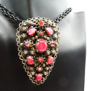 May include: Vintage-style necklace with a large, ornate pendant. The pendant features a filigree design in a bronze tone, adorned with multiple oval and round red gemstones. The necklace has a black chain.