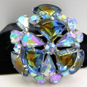 May include: A vintage brooch with a silver-tone setting, featuring large, iridescent, faceted crystals in shades of blue, green, and yellow. The brooch has a circular design with a central cluster of stones and surrounding petal-shaped crystals.