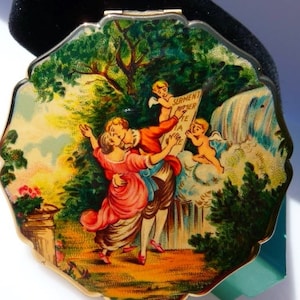 May include: A round gold-tone compact mirror featuring a colorful illustration of a couple embracing in a garden setting with two cherubs holding a sign that reads SERMENT D'AMOUR. The scene includes trees, a waterfall, and floral accents.
