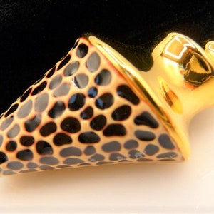 May include: A gold-colored, decorative object with a unique design. The object features a cone-shaped section with a pattern of black, irregular shapes. The edges are outlined in gold, and the top portion is also gold, with a rounded, ornate design.
