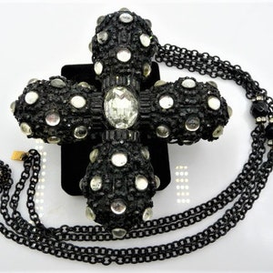 May include: A black cross pendant necklace with a large, ornate cross. The cross is encrusted with clear rhinestones and smaller, round, clear stones. The necklace has a black chain and a matching clasp. The cross measures approximately 4 inches in length.
