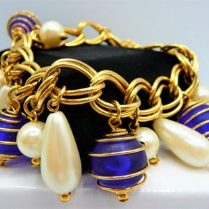 May include: Gold-tone chain bracelet with blue and gold accents. The bracelet features blue glass orbs, white teardrop-shaped beads, and round pearls. The bracelet is displayed on a black surface.
