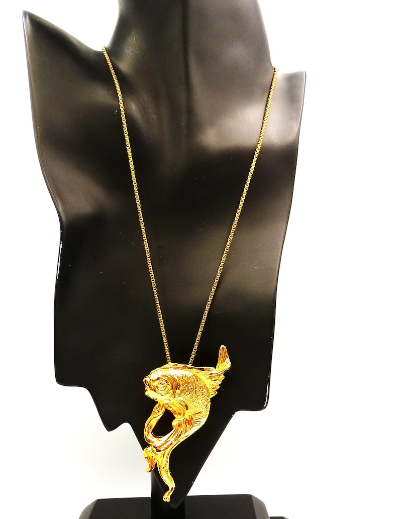 May include: A gold-colored fish pendant necklace displayed on a black mannequin. The fish pendant has detailed scales and fins, and is suspended from a delicate chain. The necklace is a statement piece, suitable for adding a touch of elegance.