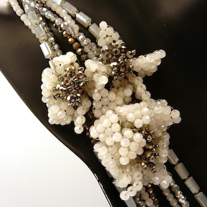 May include: A multi-strand necklace featuring white, silver, and bronze beads. The necklace has clusters of white beads and rectangular silver beads. The design is intricate and appears to be handmade, with a focus on texture and detail.