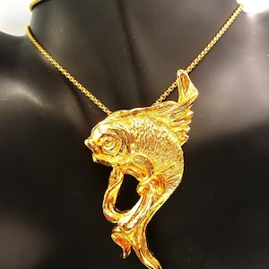 May include: A gold-colored fish pendant necklace. The pendant features a detailed fish design with flowing fins and tail. The necklace chain is a delicate gold link. The jewelry is displayed on a black mannequin.