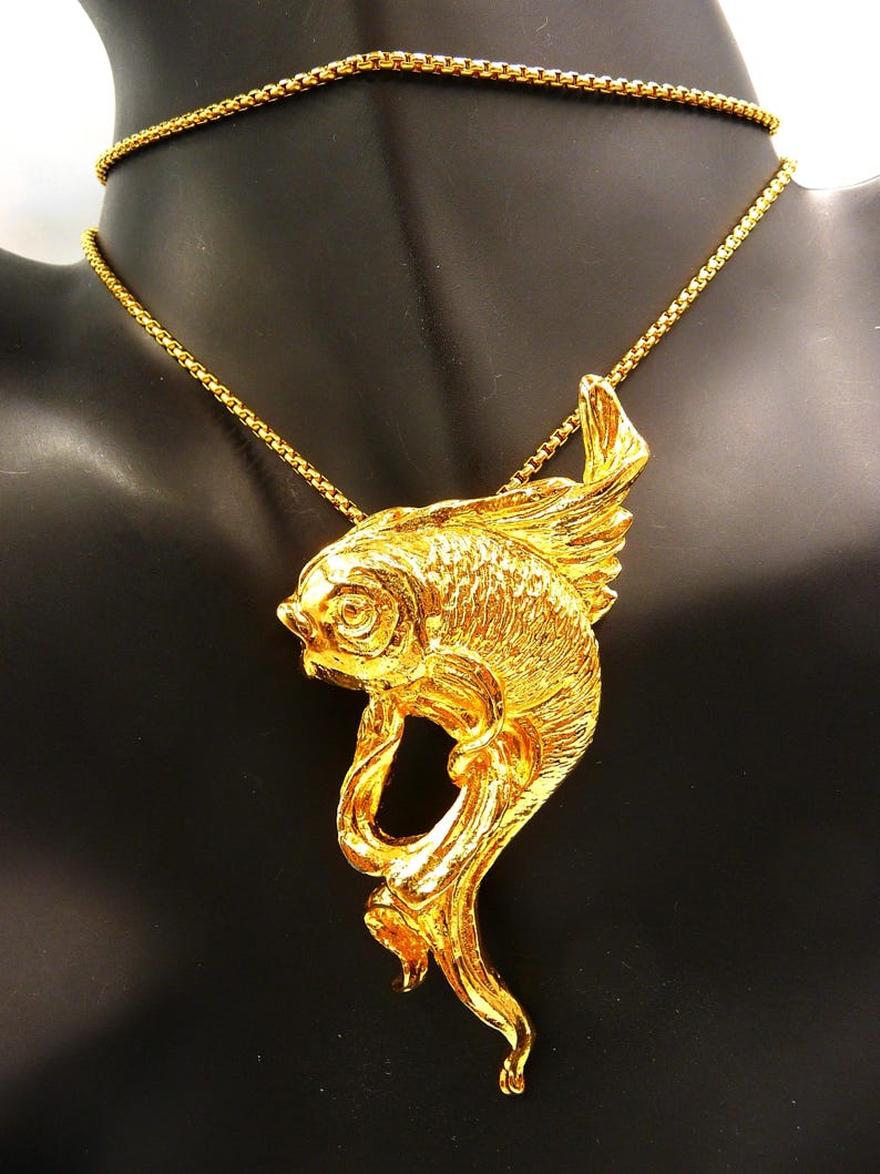 May include: A gold-colored fish pendant necklace. The pendant features a detailed fish design with flowing fins and tail. The necklace chain is a delicate gold link. The jewelry is displayed on a black mannequin.